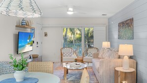 A modern living room with a glass coffee table, wicker chairs, a sofa, and a television on the wall. - Sunset Walks & Shelling Days at Seashells 9! Deeded beach access! (Sanibel)