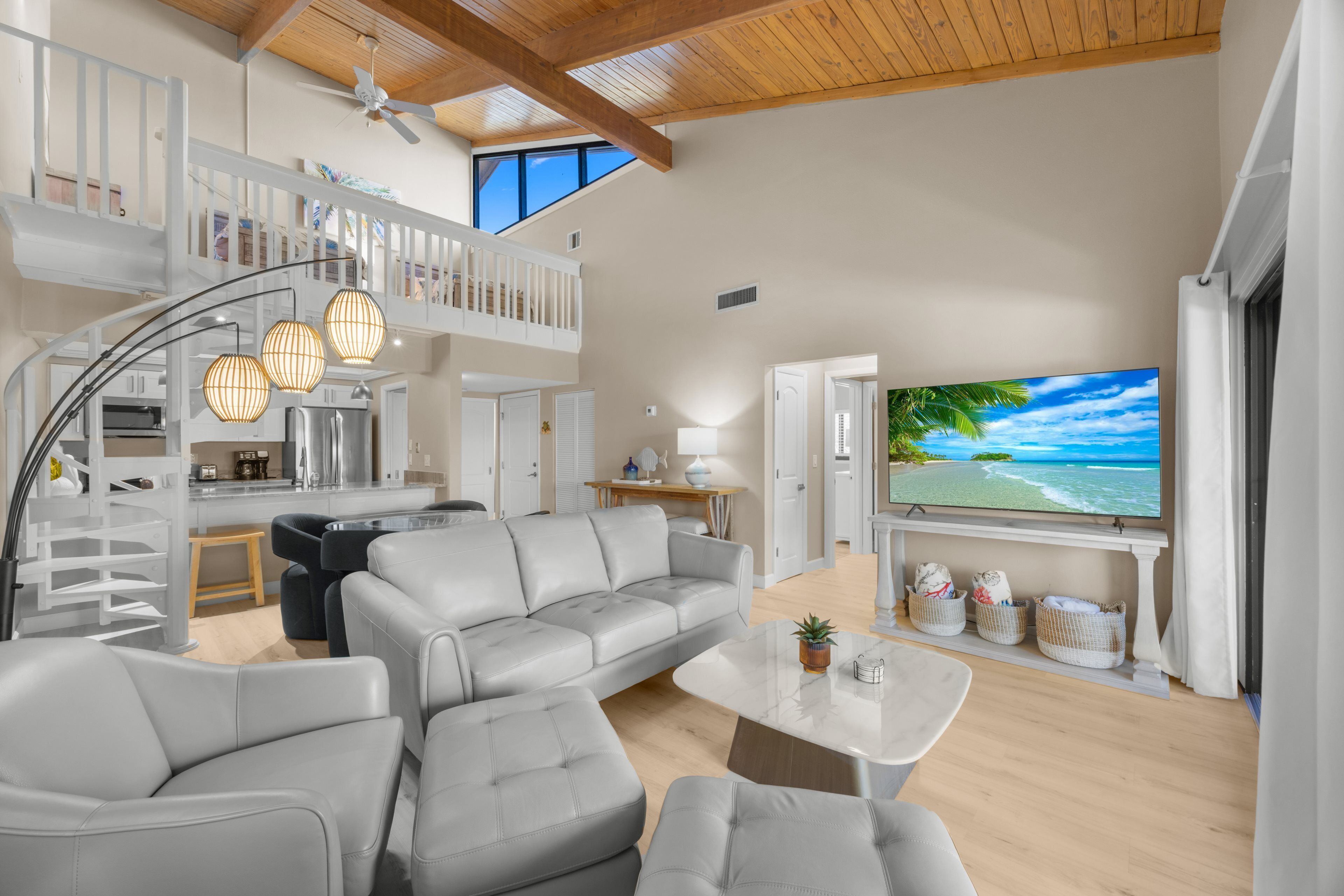 A spacious living area with a grey sofa, a coffee table, and a TV displaying a beach scene. A staircase leads to an upper level with a balcony.