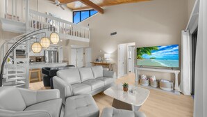 A spacious living area with a grey sofa, a coffee table, and a TV displaying a beach scene. A staircase leads to an upper level with a balcony. - Coquina Beach- Stunning Remodel-2 Bedroom- Beachside (Sanibel)