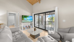 A modern living room with grey leather sofas, a marble coffee table, and a large TV screen. A sliding glass door leads to an outdoor patio with a view of palm trees and a clear blue sky. - Coquina Beach- Stunning Remodel-2 Bedroom- Beachside (Sanibel)