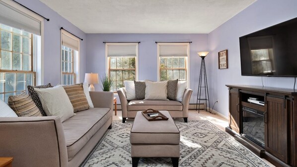 A living room with a sofa, ottoman, and a television. - Spacious 4BR Central-Berkshires Home with POOL! (Lee)