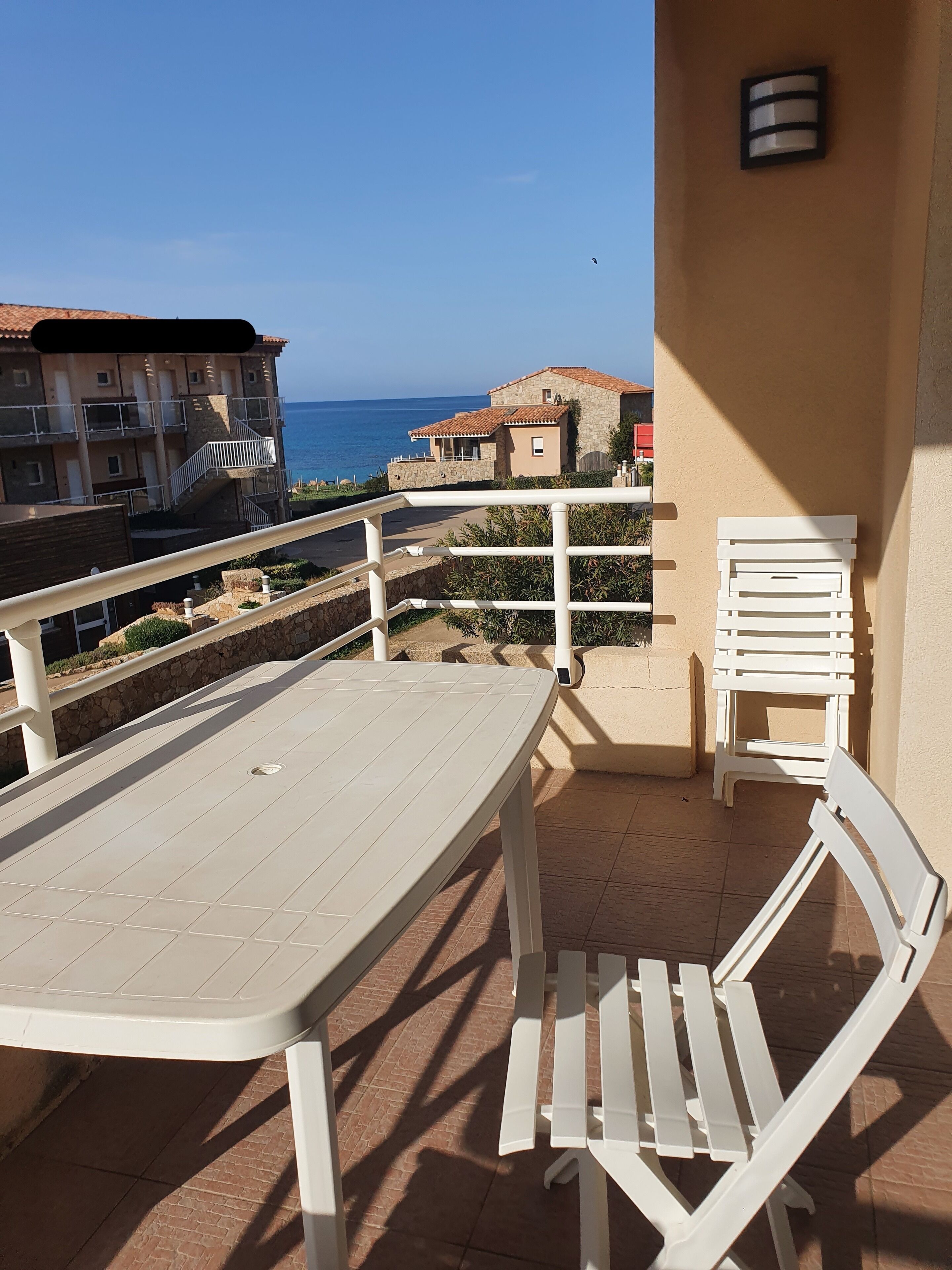 A balcony with a table and chairs, overlooking a coastal area with buildings and the sea.