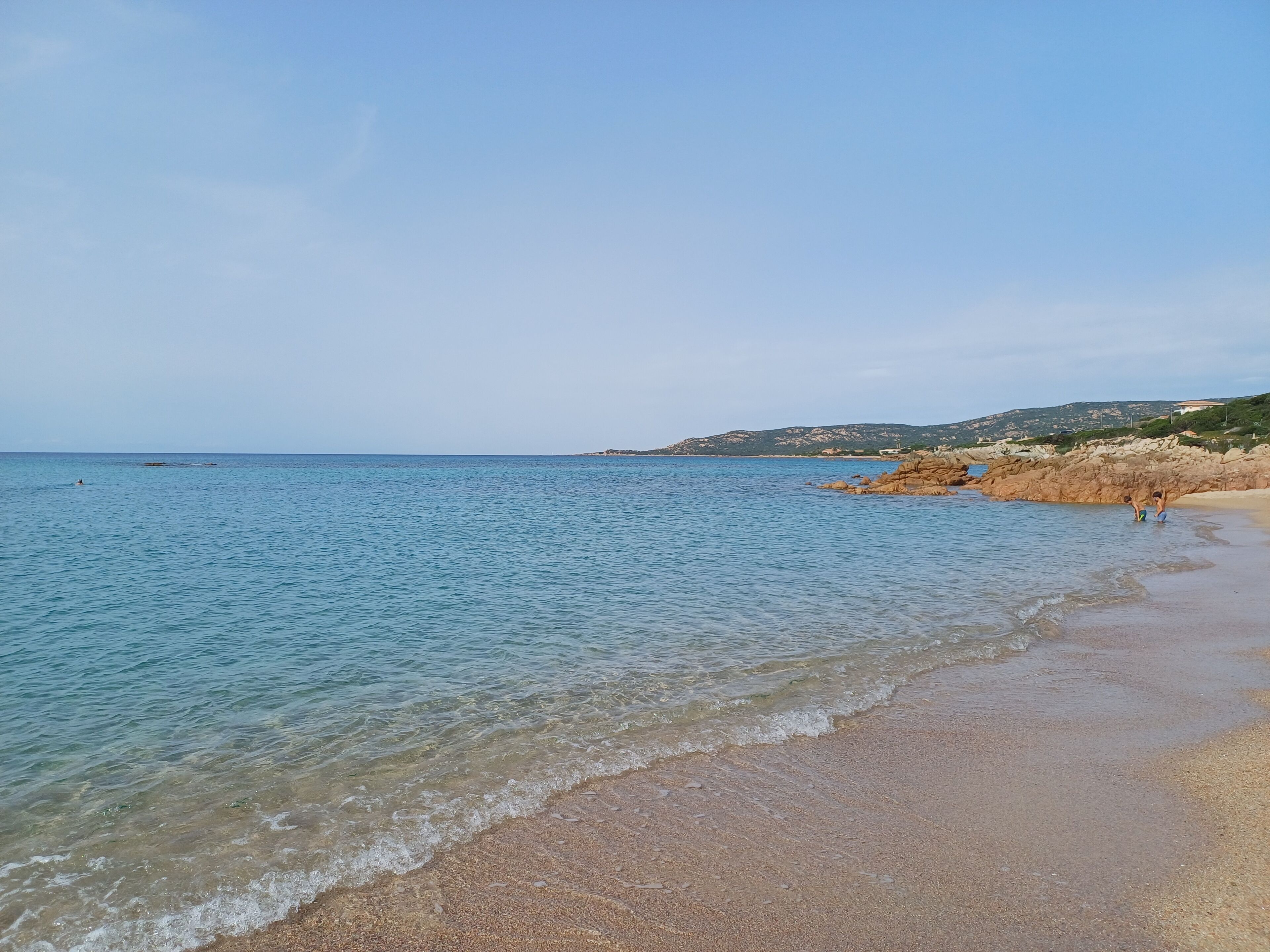 A beach with clear water, pebbly shore, and distant hills.