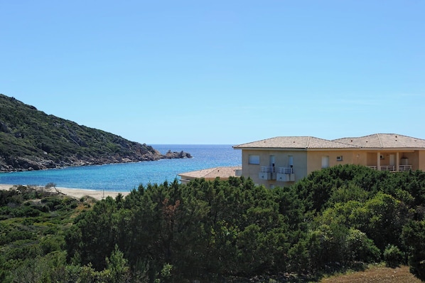 A coastal building with a balcony, surrounded by dense greenery and overlooking a clear blue sea.