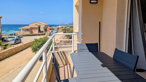 Un balcone con tavolo e sedie, affaccia su una strada costiera con auto parcheggiate e case.