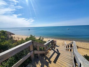 Ein hölzerner Steg führt zu einem Strand mit klarem, blauem Wasser und Booten in der Ferne.
