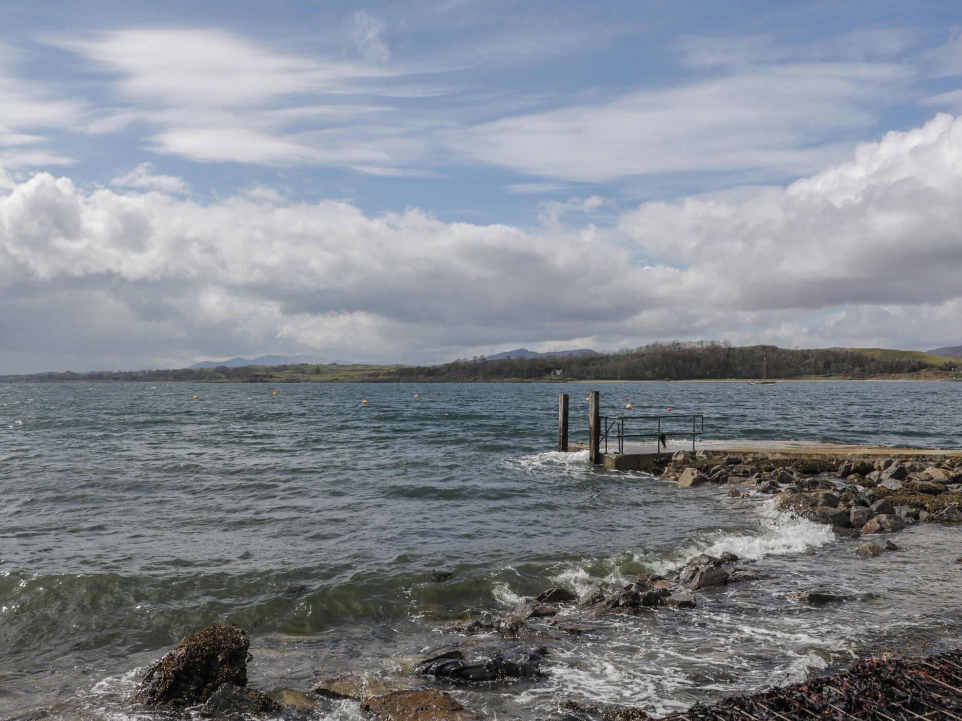 A rocky shoreline with a pier extending into calm waters, surrounded by hills and a cloudy sky.