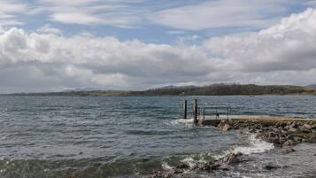 A rocky shoreline with a pier extending into calm waters, surrounded by hills and a cloudy sky.