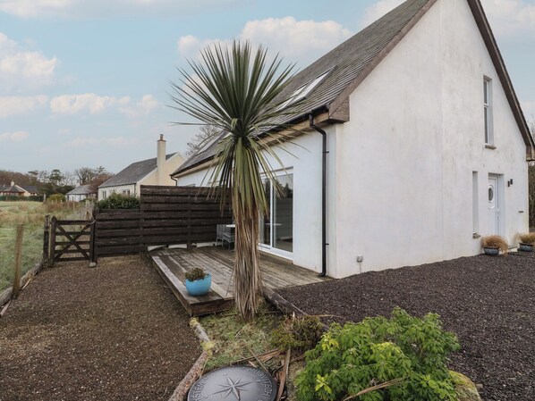 A white house with a sloped roof, a wooden fence, and a small garden area with a palm tree and potted plants. - Sealladh (Appin)