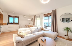 Ein modernes Wohnzimmer mit einer großen, winkelförmigen Couch, einem Essbereich mit Tisch und Stühlen sowie einer Küchenzeile mit Fenster und einer Holzarbeitsplatte.