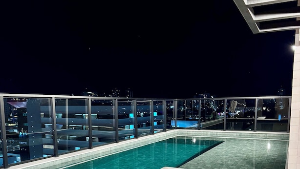 Una piscina en la azotea con agua cristalina azul, rodeada de barandillas de vidrio y vistas a la ciudad de noche.
