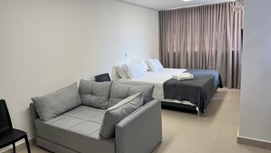 Una habitación de hotel moderna con un sofá gris, una cama con colcha oscura y una ventana con cortinas.
