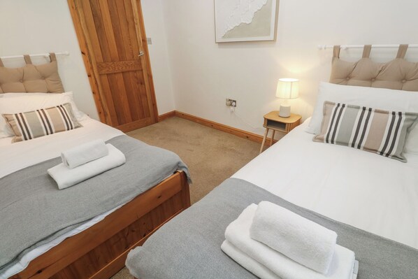 A hotel room with two beds, each with a grey bedspread and striped pillows. There is a wooden door and a small bedside table with a lamp. - Riverside Barn (Truro)