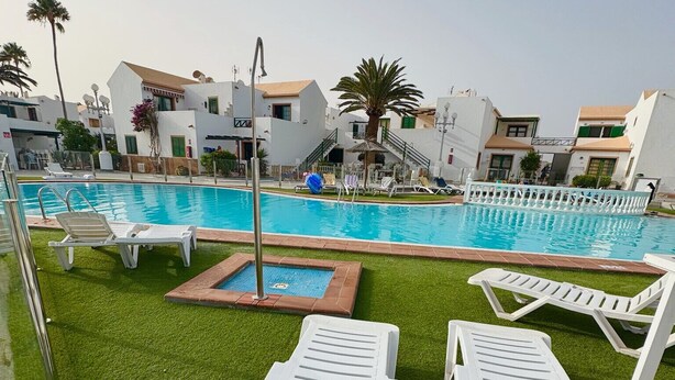 Ein Swimmingpool mit weißen Liegestühlen, einer kleinen Poolterrasse und weißen Gebäuden im Hintergrund.