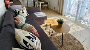 Ein modernes Wohnzimmer mit einem dunklen Sofa, einem kleinen runden Tisch mit Pflanze, einer gewebten Pendelleuchte und einer Küchenzeile mit weißen Schränken und einer Mikrowelle.
