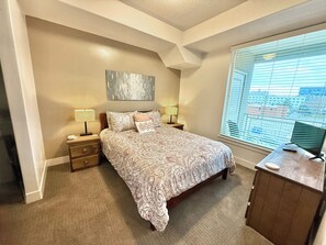 A bedroom with a large bed, two bedside tables with lamps, a dresser, and a window with blinds. - Luxury Condo in Library Square District (Salt Lake City)