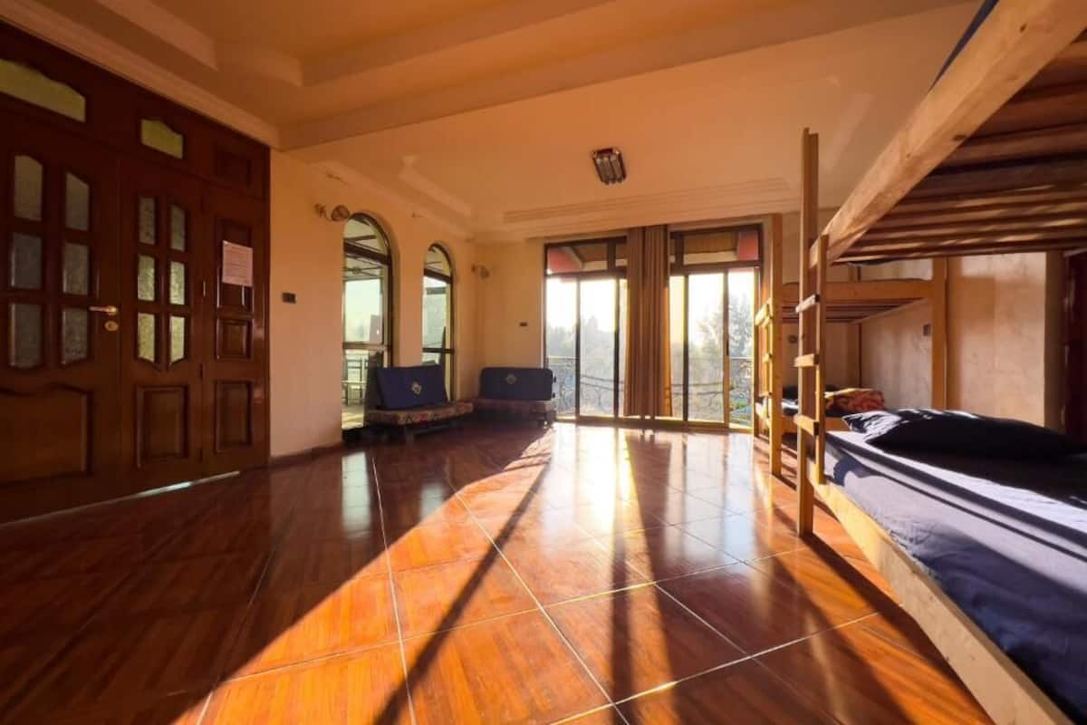 A spacious room with a wooden floor, a bunk bed, a desk with a chair, and a large window with a view of the outdoors.