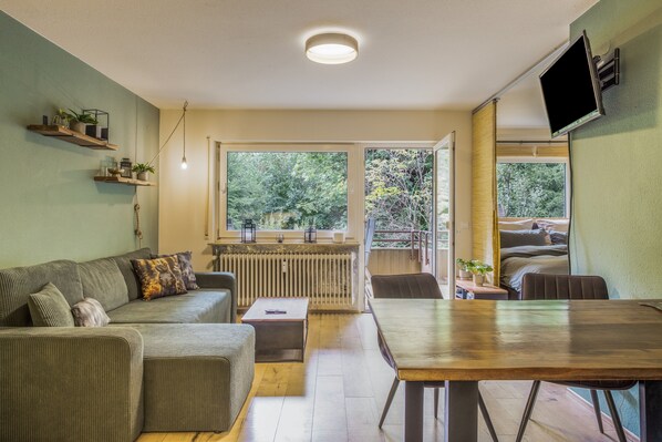A modern living room with a sofa, coffee table, and a dining table. There is a window with a view of greenery outside.