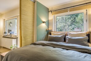 A bedroom with a bed, a bedside table, a mirror, a window with a view of trees, and a radiator.