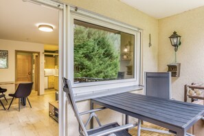 A balcony with a table and chairs, a view of a forest through a window, and a lamp on the wall.