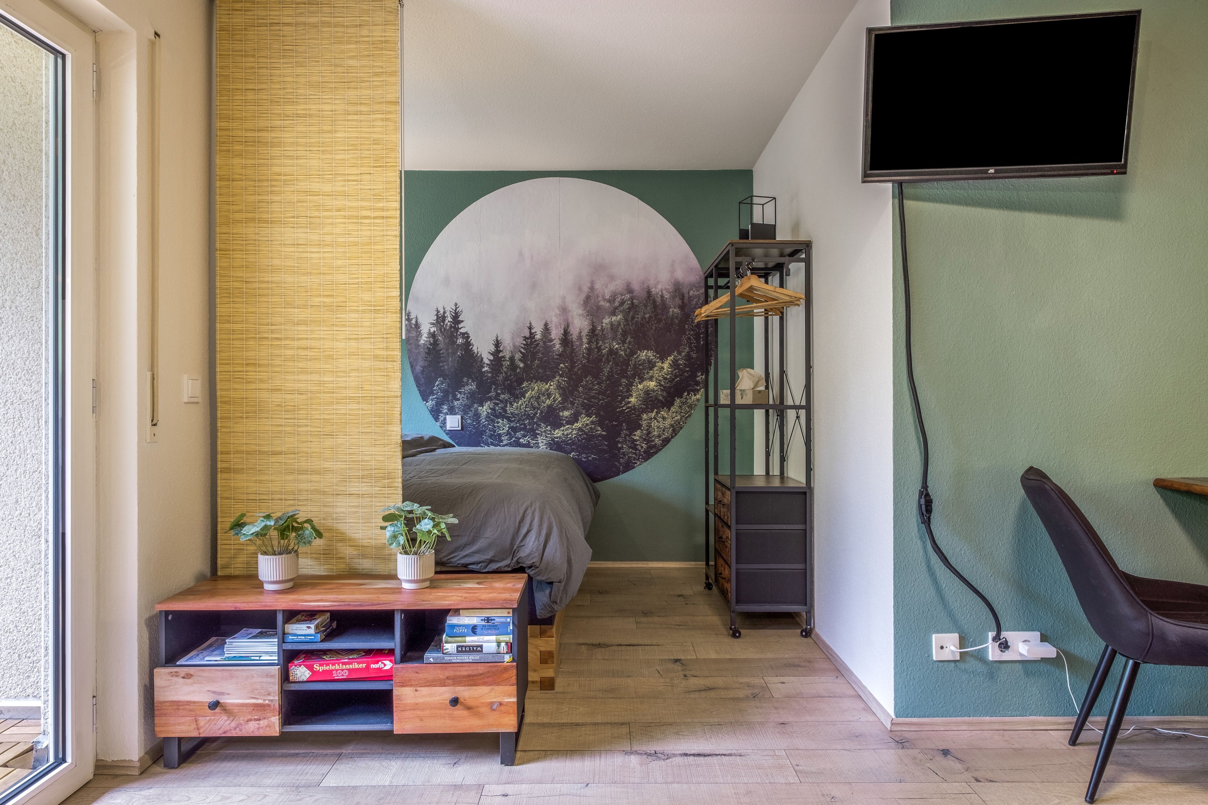 Quarto com cama, televisão, escrivaninha com livros, prateleira com objetos e parede com mural da natureza.
