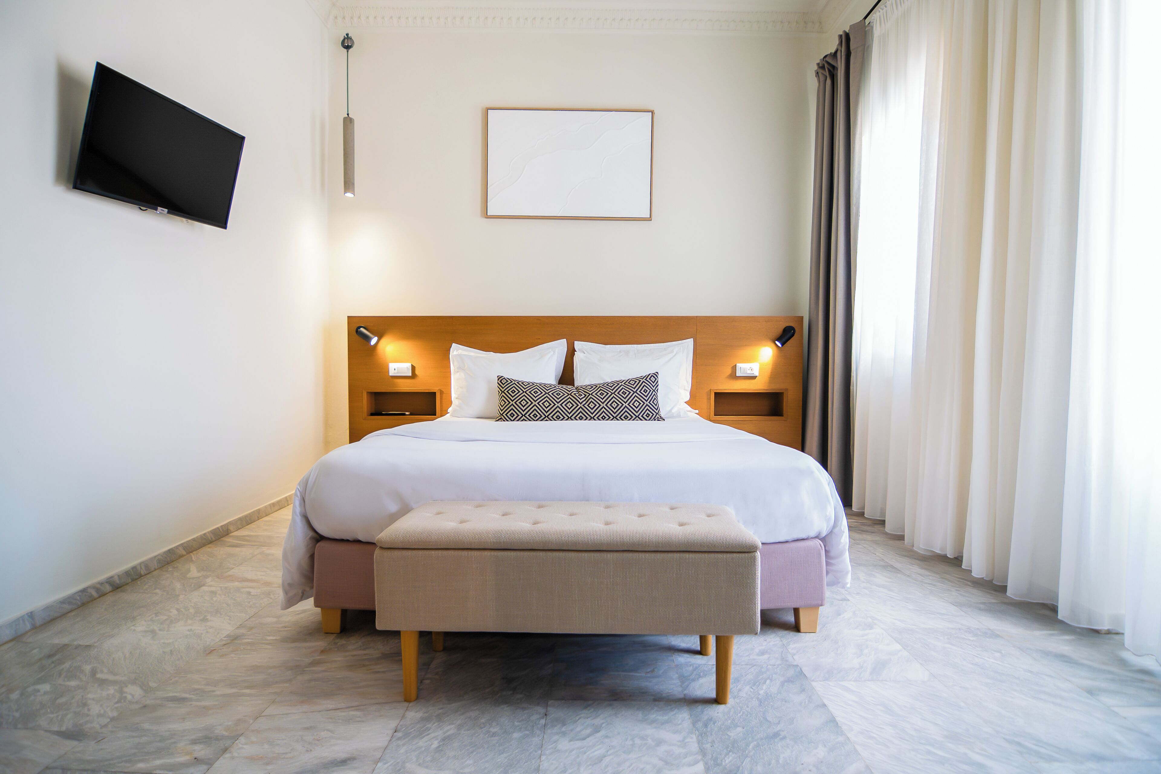 Habitación de hotel impecablemente ordenada, con una cama, lámparas de noche, un televisor de pantalla plana, un cuadro en la pared y una ventana con cortinas.