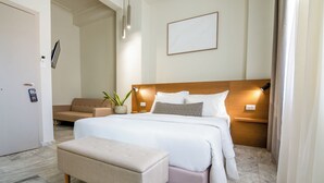 Une chambre d’hôtel avec un lit, une table de chevet, une lampe, une plante en pot et un banc.