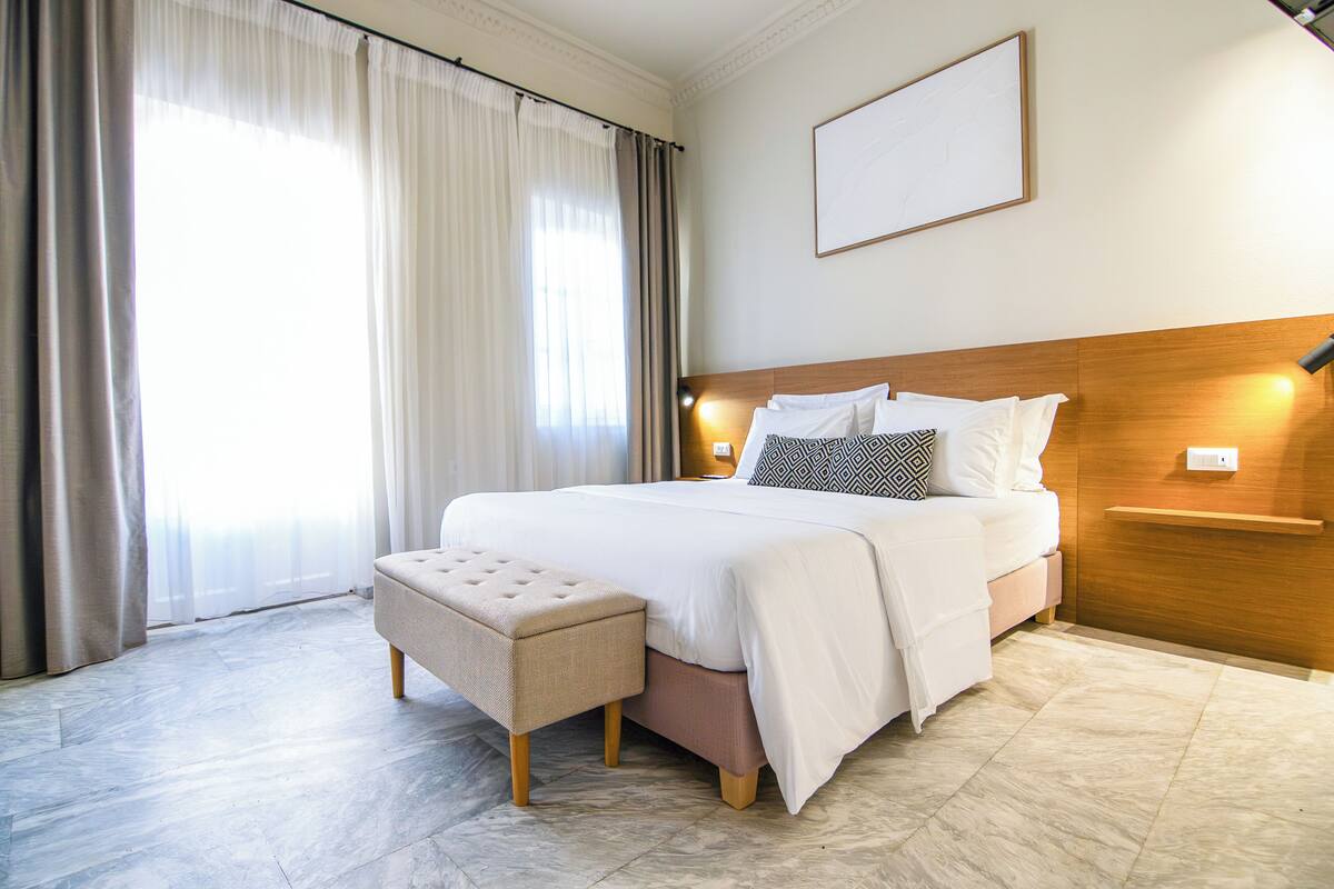 A hotel room with a large bed, a wooden headboard, a bedside table with a lamp, a bench, and a large window with curtains.