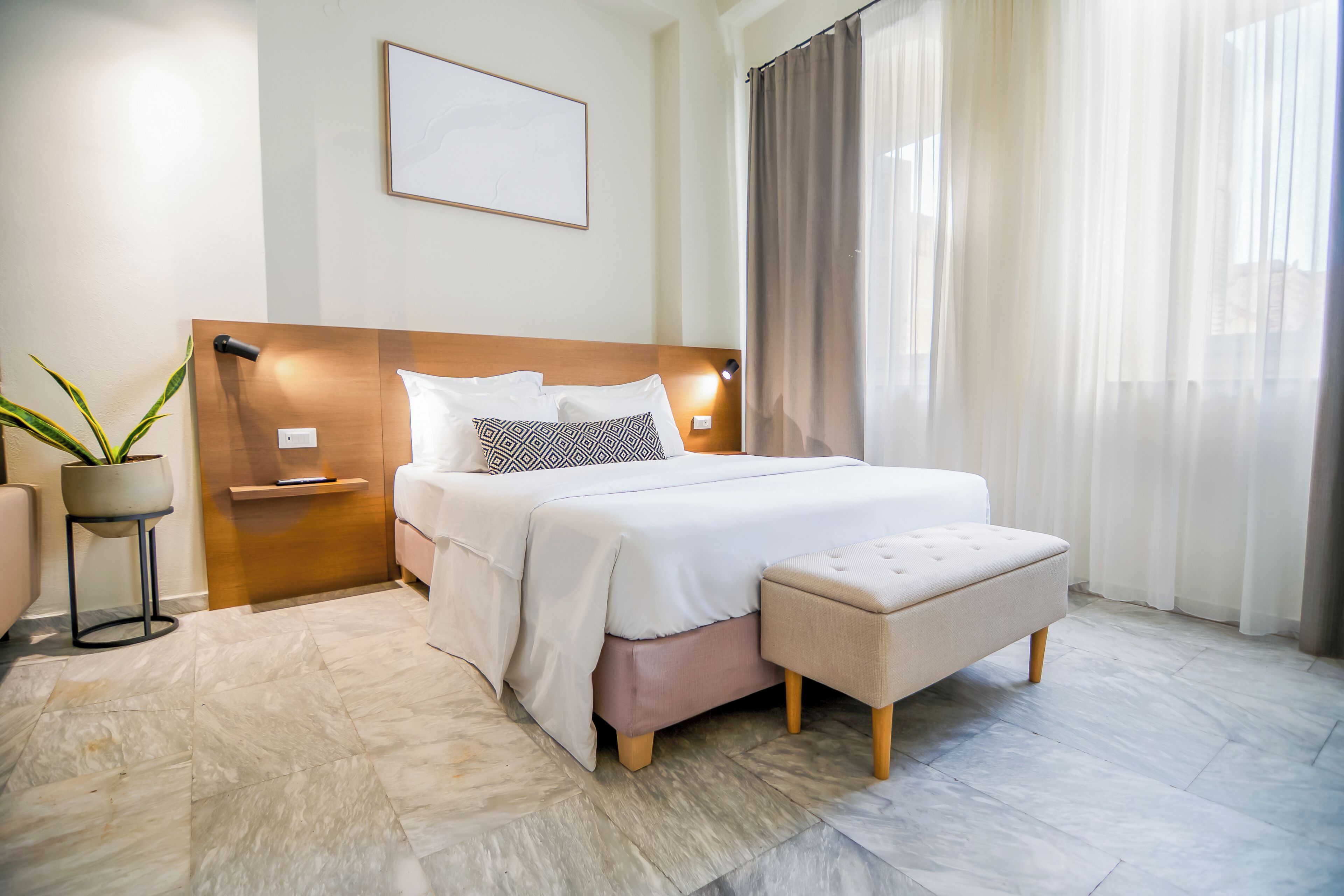 Quarto de hotel moderno com uma cama grande, mesas de cabeceira, uma planta em vaso e vista exterior através de cortinas leves.