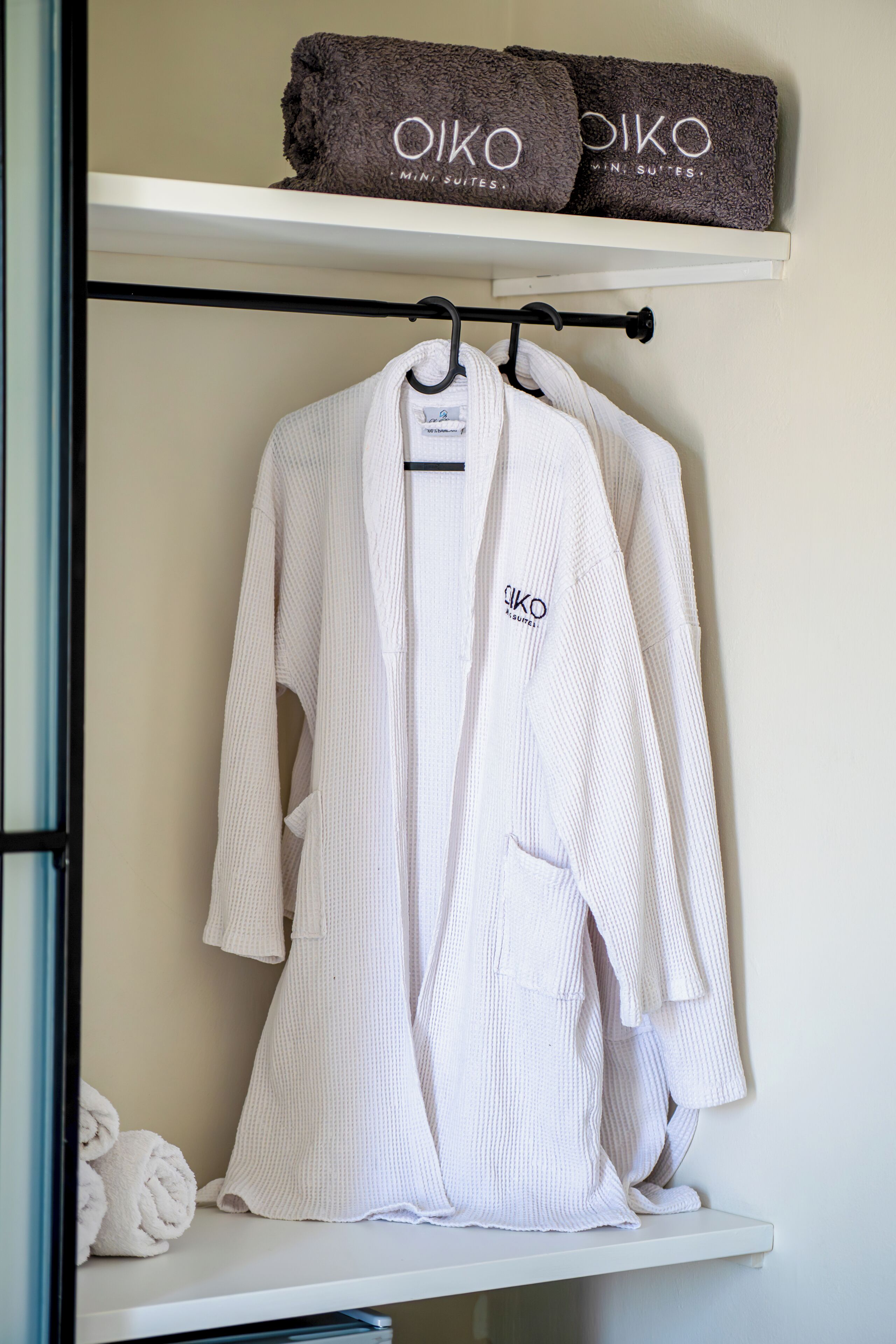 A white robe hanging on a rack, a towel, and a shelf with towels labeled 'OIKO MINI SUITES'.