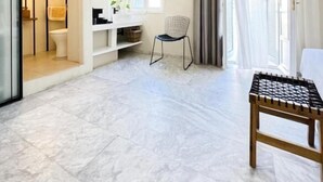 Une chambre d’hôtel moderne avec un sol en marbre, une chaise en bois, une petite table, un canapé blanc et une salle de bain visible à travers une porte ouverte.