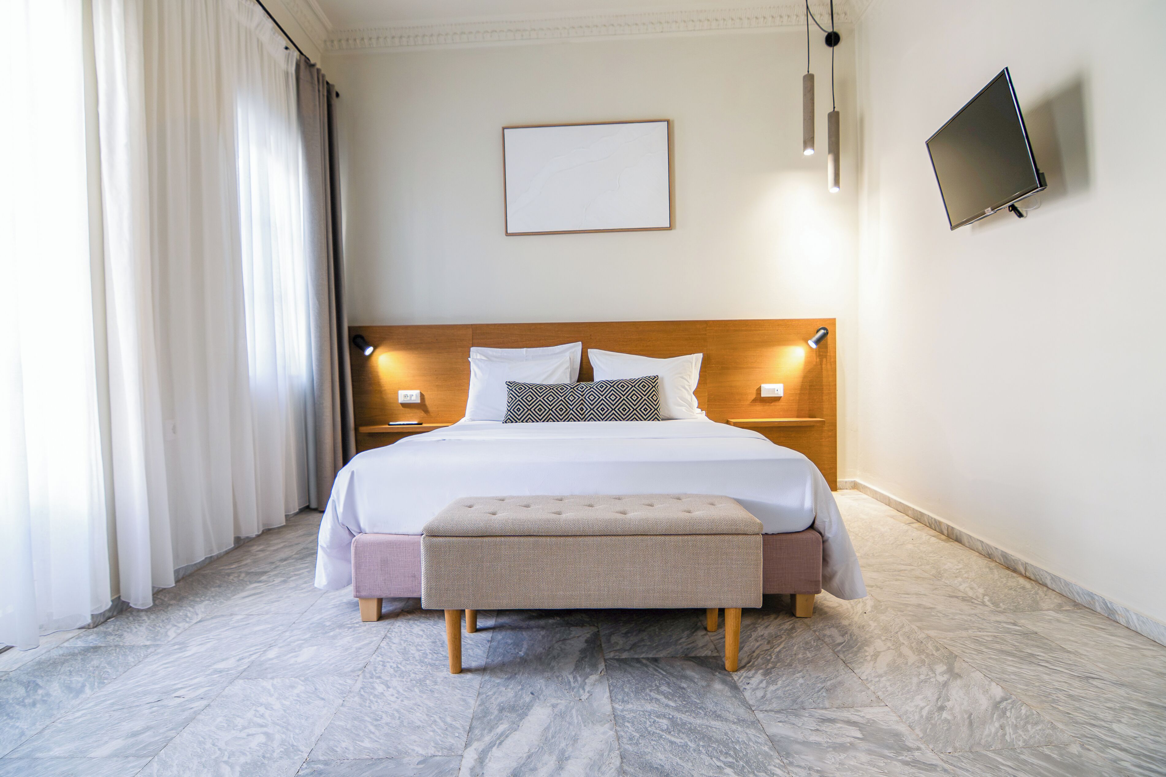 A hotel room with a bed, a flat-screen TV, a bedside table, a nightstand, and a large window with curtains.