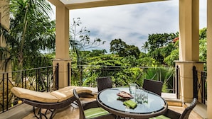 Un balcone con mobili in vimini, un tavolo in vetro e vista su una vegetazione rigogliosa.