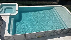 Ein rechteckiger Swimmingpool mit einem Whirlpoolbereich, umgeben von einer Betonplattform und einem Holzzaun.