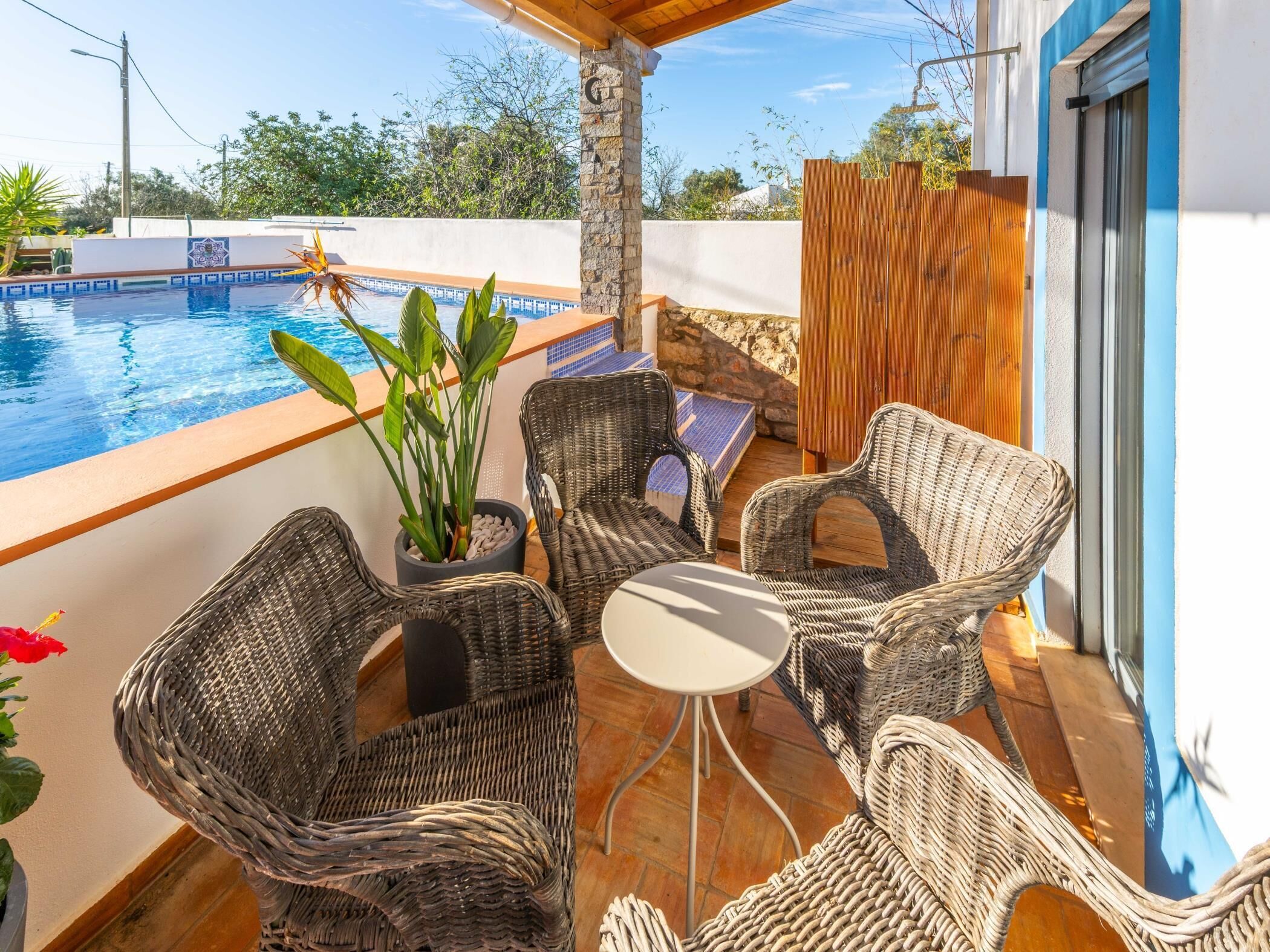 Un área junto a la piscina con sillas de mimbre y una mesa, rodeada por una terraza de madera y un muro de piedra.