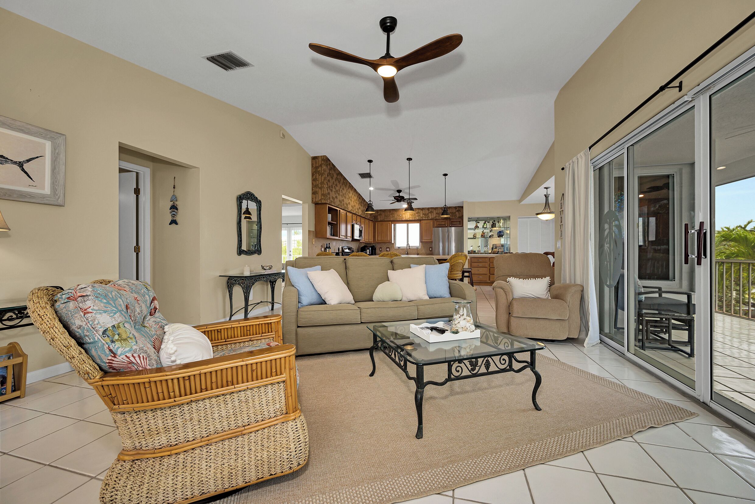 A spacious living area with a large sofa, a wicker chair, and a glass coffee table. There is a ceiling fan, a kitchen in the background, and a sliding glass door leading to an outdoor area.