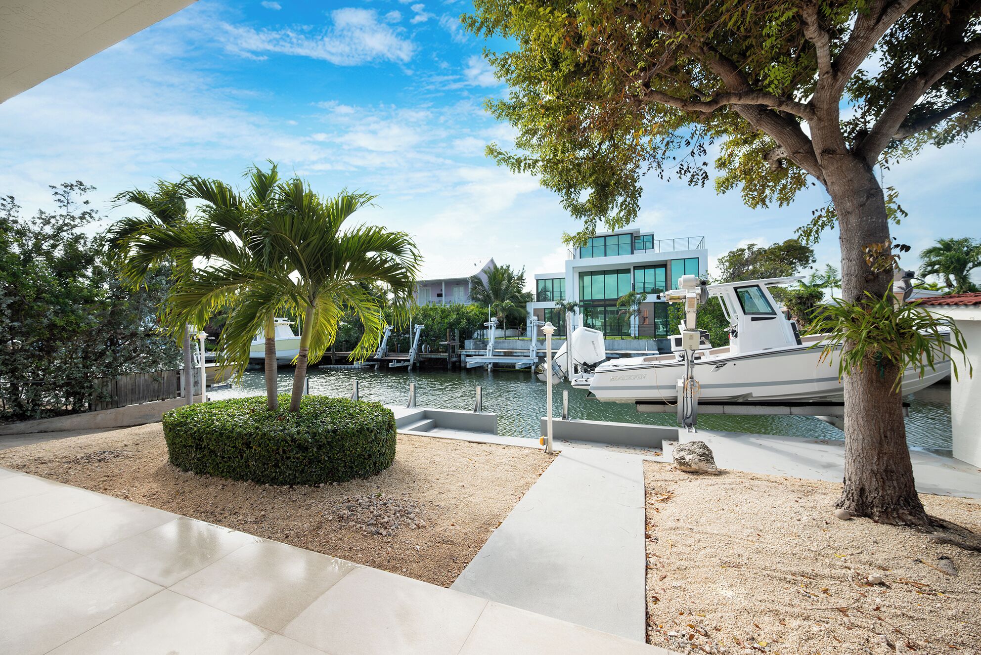 A marina with boats docked, a modern building with large windows, and a well-maintained landscaped area with palm trees and a hedge.