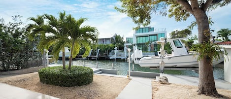 A marina with boats docked, a modern building with large windows, and a well-maintained landscaped area with palm trees and a hedge.