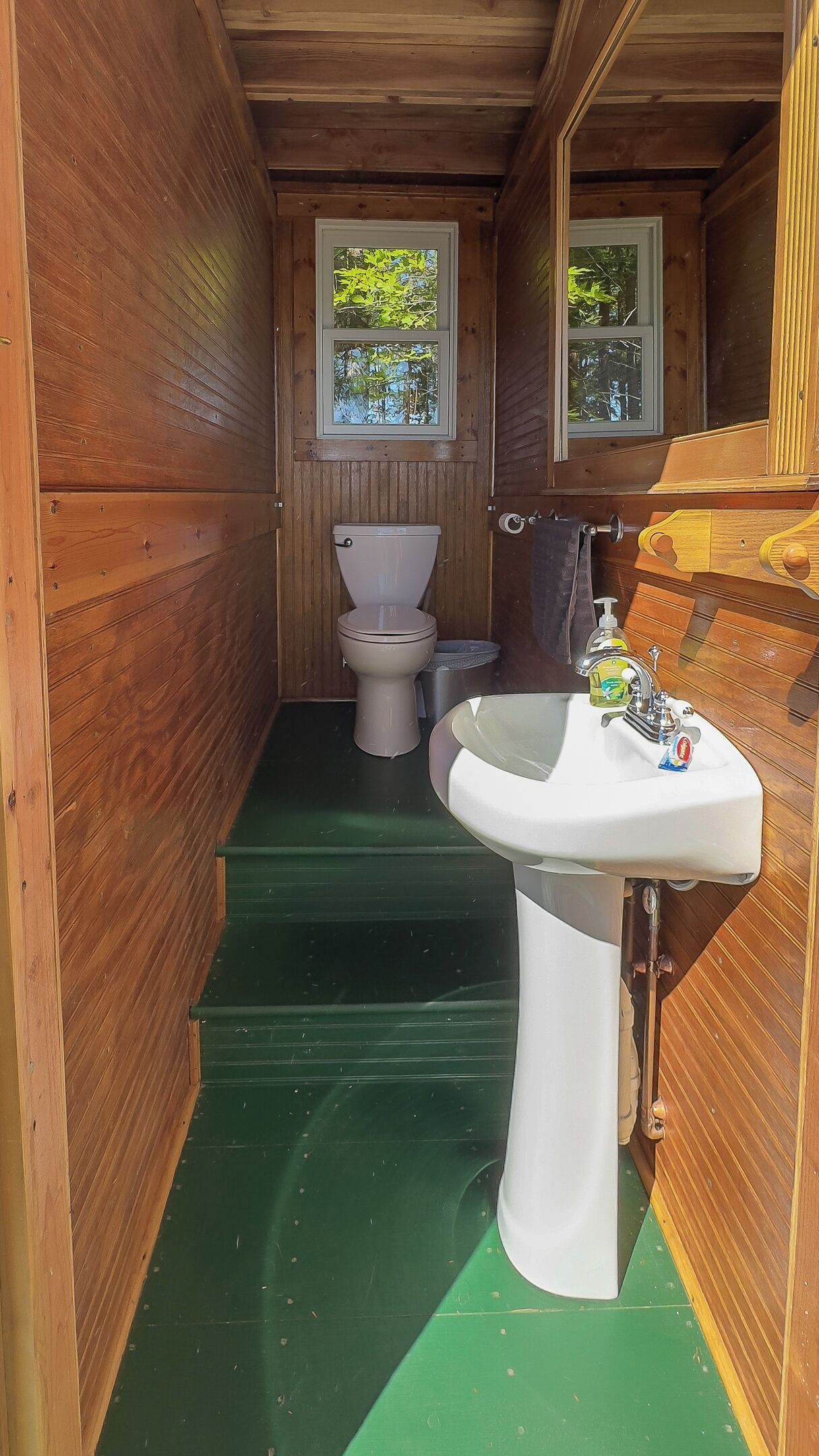 A compact wooden bathroom with a toilet, sink, and two windows.