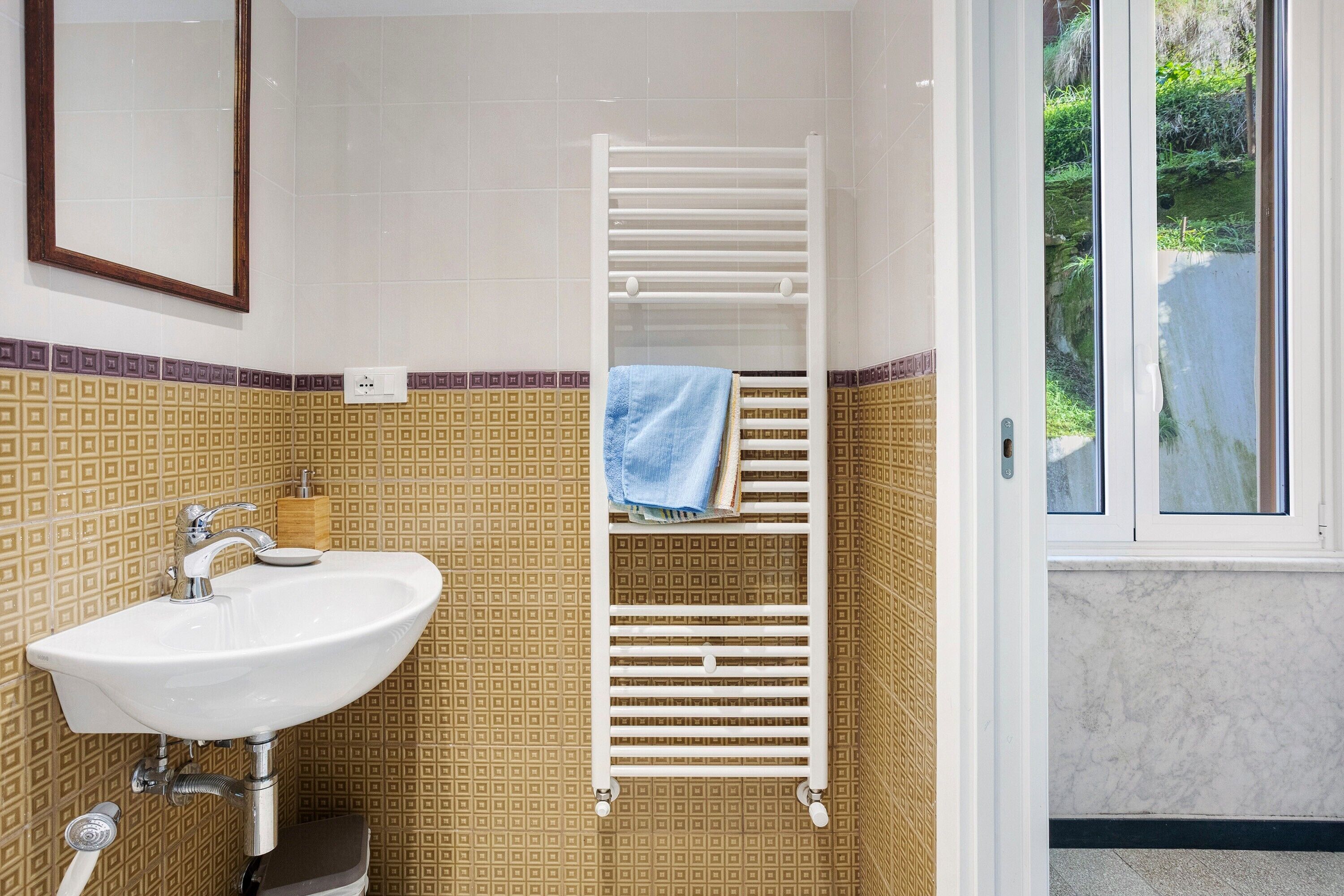 A bathroom with a white sink, a towel rack, and a window with a view of greenery.