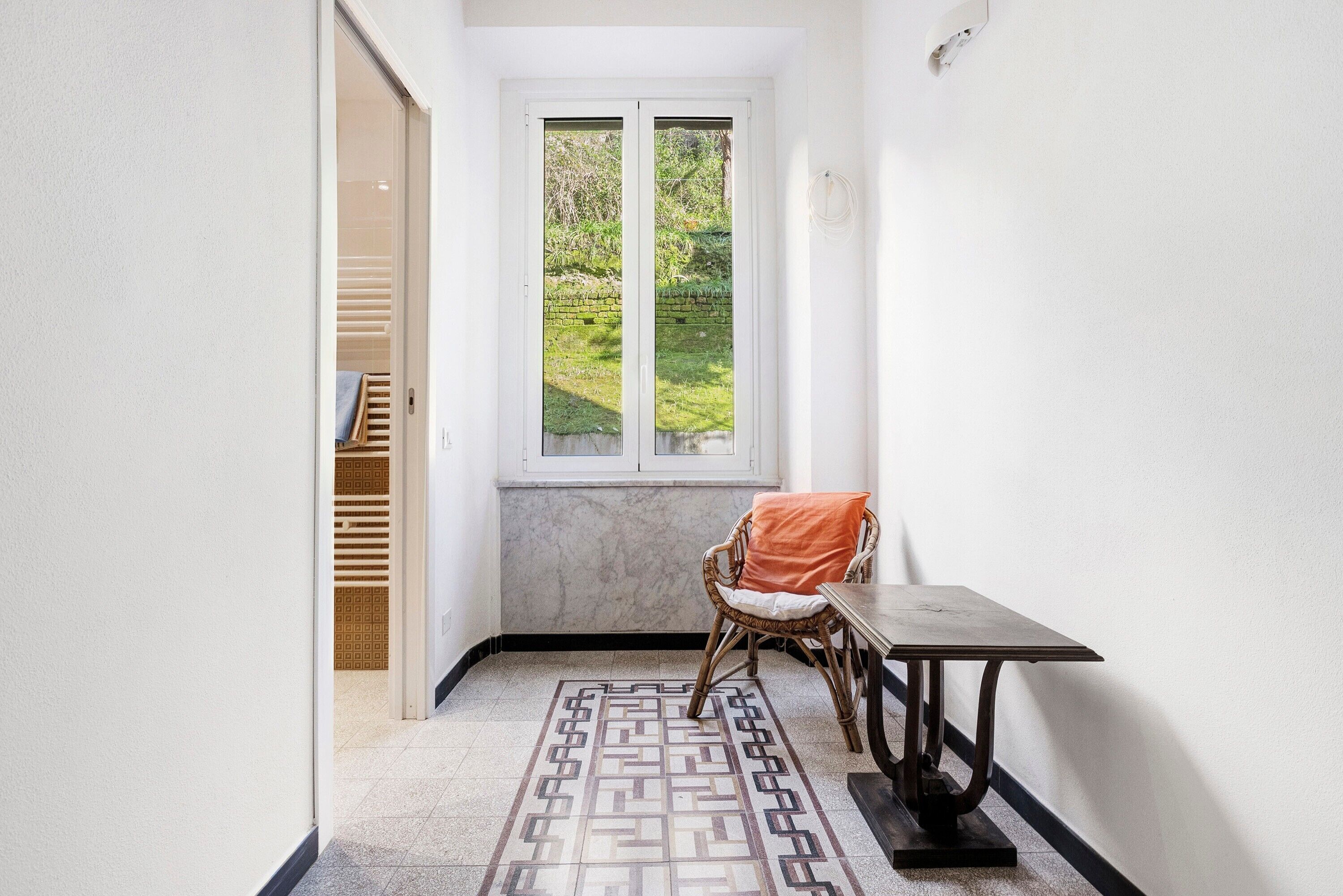 Un pasillo estrecho con un tapete estampado, una silla de mimbre con cojín, una mesita y una ventana con vistas a zonas verdes.