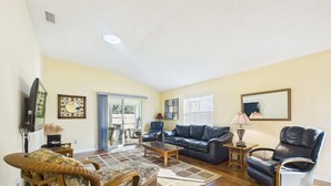 A spacious living room with a large sofa, a coffee table, and a wicker chair. There is a television and a lamp on the wall. - Cozy Courtyard Villa with Pool in De La Vista (The Villages)