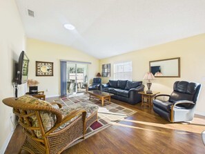 A spacious living room with a large sofa, a coffee table, and a wicker chair. There is a television and a lamp on the wall. - Cozy Courtyard Villa with Pool in De La Vista (The Villages)