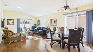 A spacious living area with a dark leather sofa, wicker chairs, a glass dining table, and a ceiling fan. - Cozy Courtyard Villa with Pool in De La Vista (The Villages)
