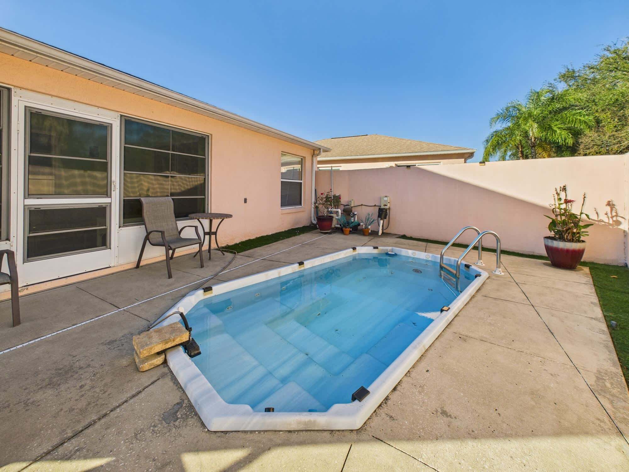 Piscina rectangular con una escalera en un lateral, rodeada por un patio de hormigón. Hay dos sillas y una mesita cerca de la piscina, y una planta en maceta a la derecha.