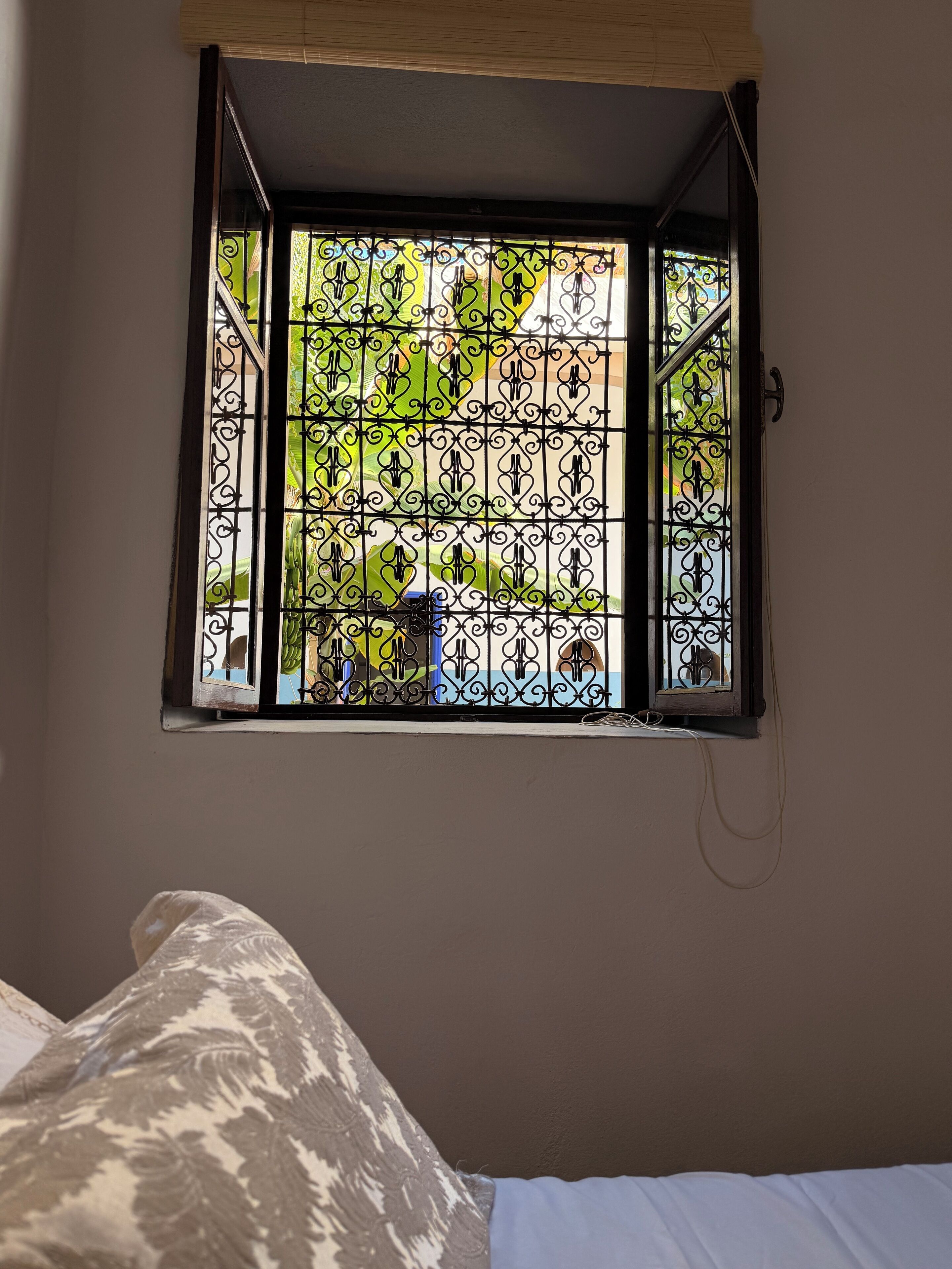 Ein Fenster mit schmiedeeisernen Ziergittern, ein Bett mit gemusterten Betttextilien und eine Wandleuchte.