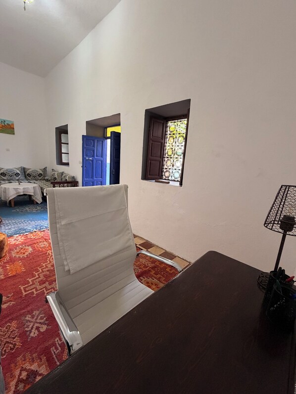 A room with a white chair, a dark wooden table, a patterned rug, a blue door, and a window with a decorative grille.