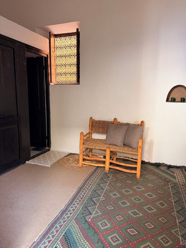 A room with a wooden rocking chair, a patterned rug, a small shelf with two green cups, and a window with decorative iron bars.
