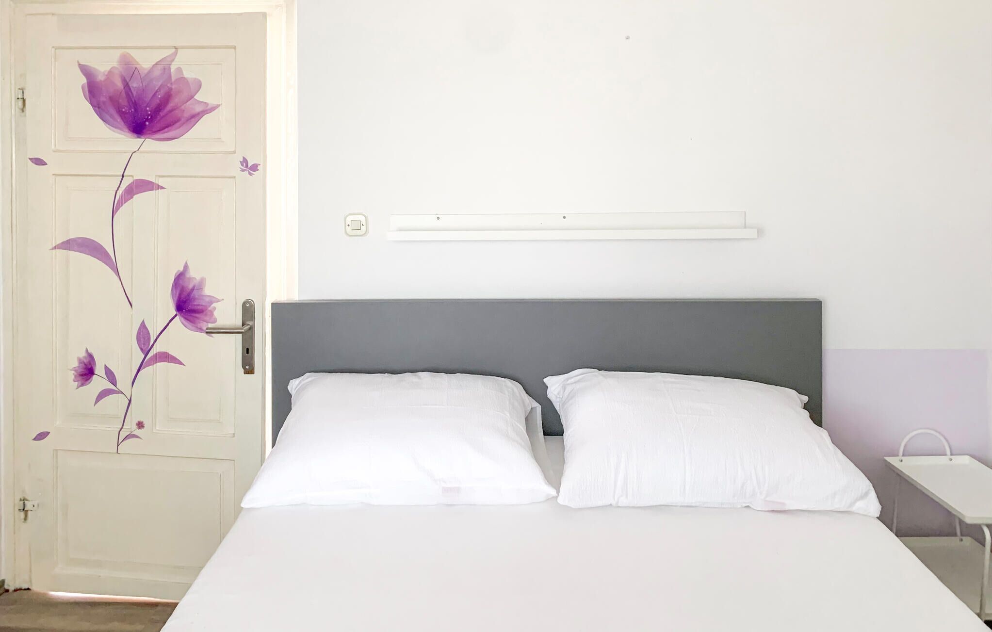 A bedroom with a bed, two white pillows, a grey headboard, a nightstand, and a wall-mounted light fixture.