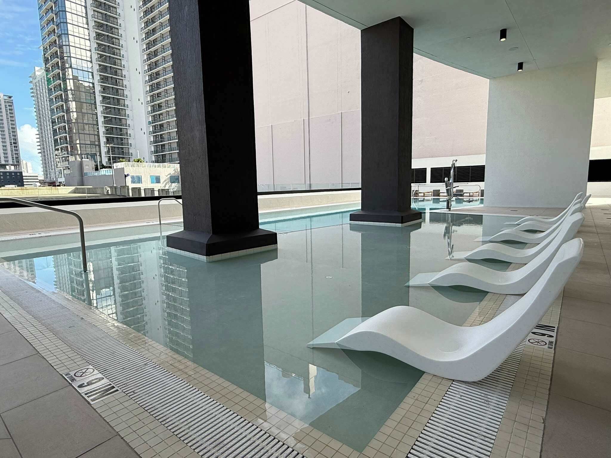 A modern indoor pool with clear glass walls, black pillars, and white seating.